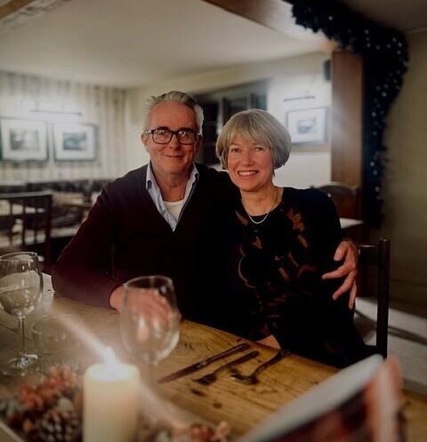 Geoff is sat smiling facing the camera in a rustic restaurant with his left arm wrapped around his wife. They are sat down behind a wooden table with two wine glasses and cutlery on top. Geoff is wearing his black long-sleeved polo top that is slightly unzipped in front.