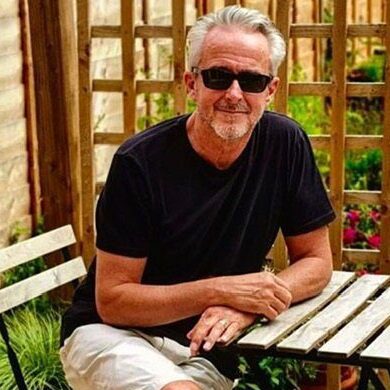 Geoff is sat outside on the patio surrounded by greenery smiling at the camera. He has his legs crossed and hands overlapping on one another while placed on the wooden table. He is wearing black sunglasses, black t shirt with beige shorts.
