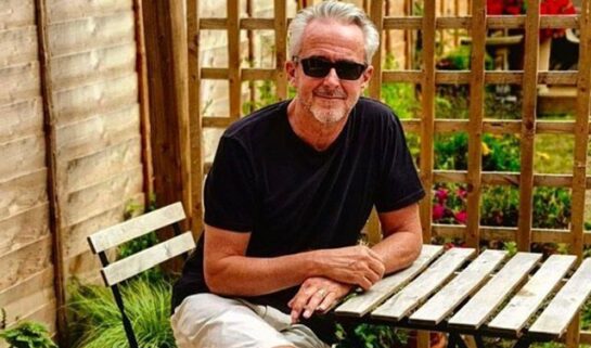 Geoff is sat outside on the patio surrounded by greenery smiling at the camera. He has his legs crossed and hands overlapping on one another while placed on the wooden table. He is wearing black sunglasses, black t shirt with beige shorts.