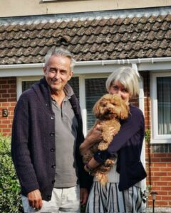 Geoff is wearing a dark blue cardigan with a grey polo top smiling at the camera. His wife is stood next to him holding their chestnut cockapoo. They are stood in front of the back of their red brick layered house.