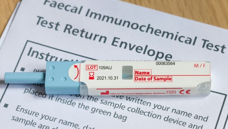 An unlabelled faecal immunochemical test sample tube lies on top of a sheet of paper that reads faecal immunochemical test test return envelope with instructions on top of a wooden table.
