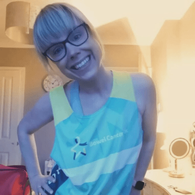 Rachel is grinning at the camera in a room in her house. She has her right hand on her hip and she is wearing the blue Bowel Cancer UK vest.
