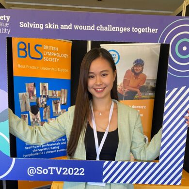 Dove is at a conference wearing a pastel green suit and holding a large frame in front of her that is from the society of tissue viability. Her upper half of her body is inside the frame. Behind her is an large orange banner and table labelled as the British Lymphology Society.