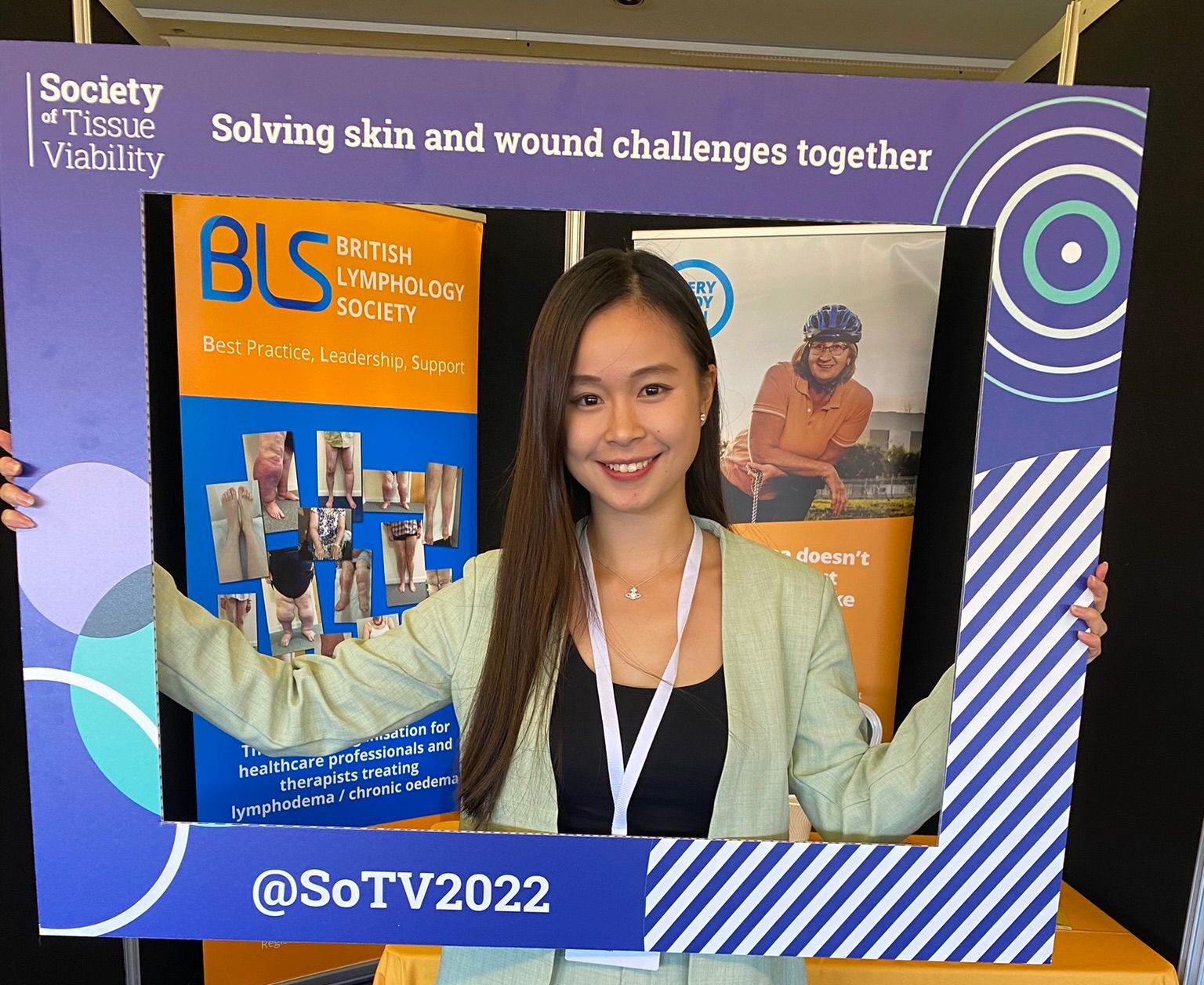 Dove is at a conference wearing a pastel green suit and holding a large frame in front of her that is from the society of tissue viability. Her upper half of her body is inside the frame. Behind her is an large orange banner and table labelled as the British Lymphology Society.