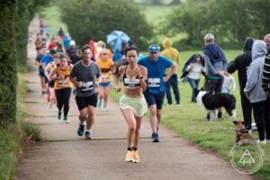 Dove is seen in a race running uphill. She is wearing a sports bra attached to a running number in front with pastel green running shorts and bright orange trainers. She is amongst many other runners with people standing beside the track and observing on the grass. 