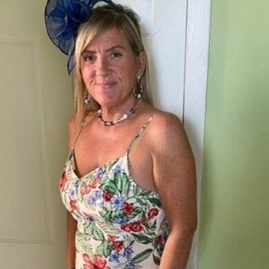 Lorna is stood in front a white wooden door and green walls. She is smiling at the camera with her blonde hair resting on her shoulder. She is wearing a long white summery floral dress and a navy flower fascinator hat.