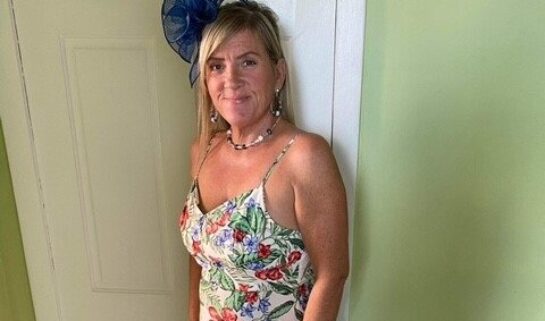 Lorna is stood in front a white wooden door and green walls. She is smiling at the camera with her blonde hair resting on her shoulder. She is wearing a long white summery floral dress and a navy flower fascinator hat.