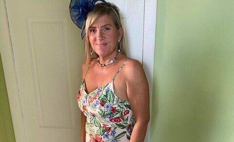 Lorna is stood in front a white wooden door and green walls. She is smiling at the camera with her blonde hair resting on her shoulder. She is wearing a long white summery floral dress and a navy flower fascinator hat.