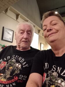 Diane is taking a selfie with her friend with a grin on her face. They wear black Mad Sunday 2022 Isle of Man t-shirts and are indoors with high ceilings and white walls.