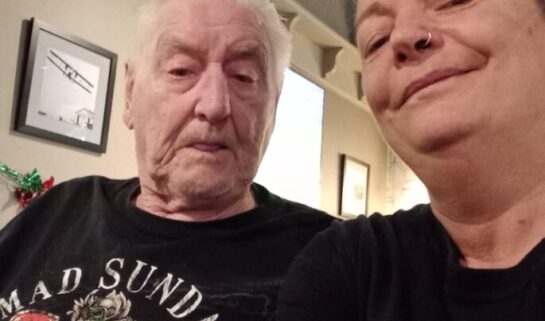Diane is taking a selfie with her friend with a grin on her face. They wear black Mad Sunday 2022 Isle of Man t-shirts and are indoors with high ceilings and white walls.