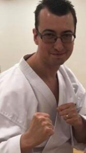 Matt is wearing his white taekwondo outfit, also known as a dobok and making two fists into a fighting gesture while smiling at the camera. 