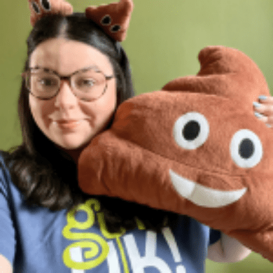 Jade has shoulder length dark hair and round glasses. She is wearing a Guts UK t-shirt and poo head boppers. She is holding up a brown poo cushion with eyes and a smile. She is smiling at the camera. The wall behind her is green.