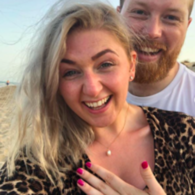 Alicia is taking a selfie with her partner at the beach. She has a big smile on her face and has placed her left hand on her chest. A wedding ring is seen.