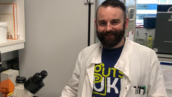 Dr. Daniel Patten is stood smiling at the camera inside a lab and wearing the Guts UK t-shirt under his white lab coat. He has his right hand placed besides a microscope on top of a white bench.