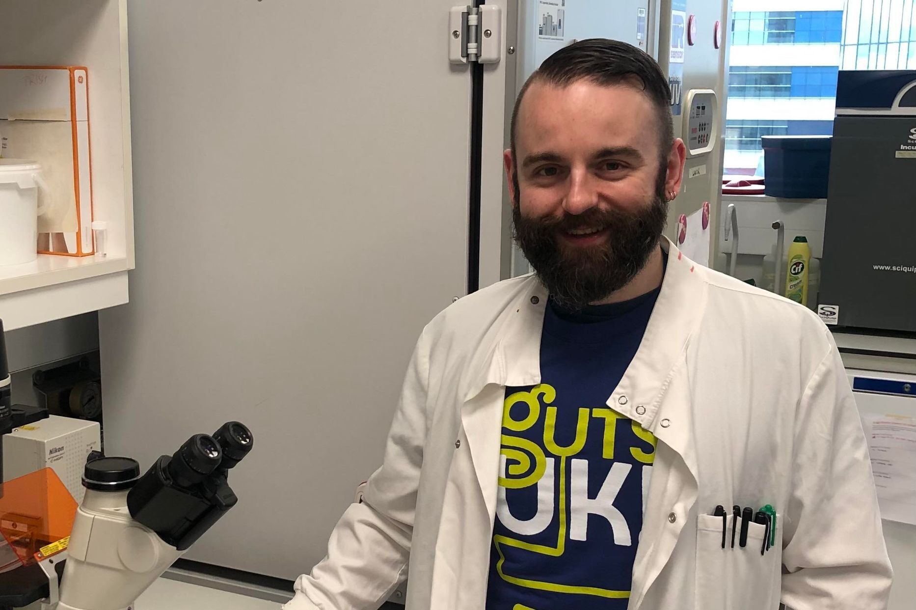 Dr. Daniel Patten is stood smiling at the camera inside a lab and wearing the Guts UK t-shirt under his white lab coat. He has his right hand placed besides a microscope on top of a white bench. 