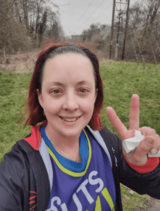 Hannah is taking a selfie outdoors in her Guts UK vest top. She is smiling at the camera and has her fingers in a 'V' shape. 
