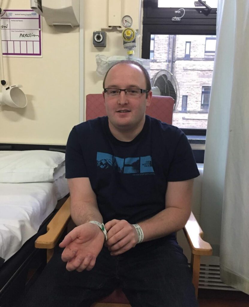 Ross awaiting surgery in hospital. He is wearing a navy t-shirt and jeans, glasses and is smiling nervously at the camera, whilst holding his hospital admission bracelet on his arm.