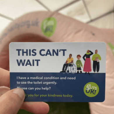 A hand is holding one of Guts UK's 'This can't wait' cards, which is a wallet sized card that helps people gain access to public toilets urgently. The card is white and blue, with Guts UK's green circular logo and an illustration of four people standing next to each other.