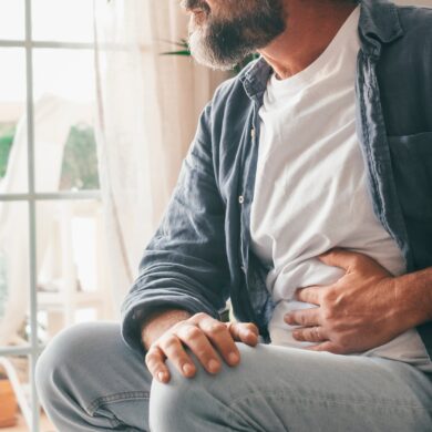 A man is sat down. He is holding his stomach in pain. He is wearing a white shirt, light blue jeans and a blue button shirt.