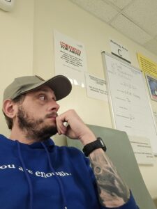 Ian is sat in a hospital chair and looking down at the corridor with his hand on his chin. He is wearing a blue jumper and a green hat.