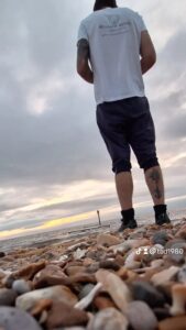 Ian is wearing a white top and gray trousers rolled up on a pebble beach. His back is in the photo and a light tower is seen in the distance in the gray and cloudy skies.