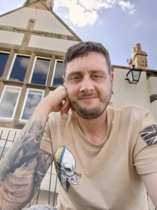 Ian is sat outdoors in front of a building. He is smiling at the camera wearing a beige t-shirt with his right hand pushed against his face. 