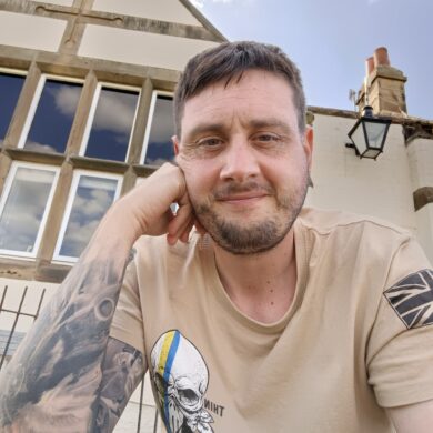 Ian is sat outdoors in front of a building. He is smiling at the camera wearing a beige t-shirt with his right hand pushed against his face.