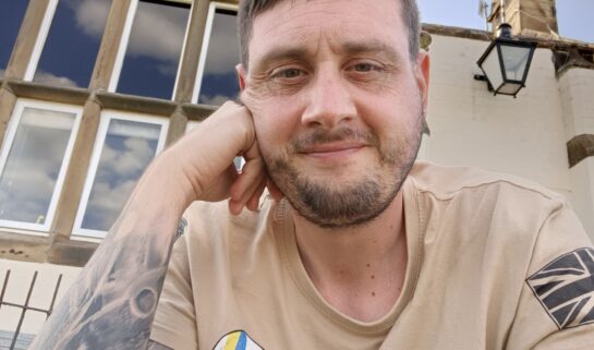 Ian is sat outdoors in front of a building. He is smiling at the camera wearing a beige t-shirt with his right hand pushed against his face.