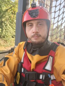 Ian is taking a selfie outdoors and is wearing a orange lifeguard uniform with a red reflective vest and red helmet.