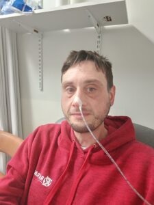 Ian is sat on a hospital chair wearing a red jumper. He has a plastic tube going through his nose. 