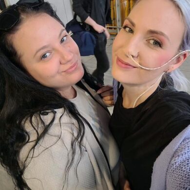 Leanne is taking a selfie with her friend and they are both smiling at the camera. Leanne is wearing a black top with a purple cardigan and he friend is wearing a beige jumper. Leanne has a tube passing through into nose.