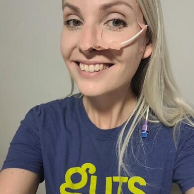 Leanne has long blonde hair, which she is wearing down. She is smiling at the camera. She has a feeding tube running into her nose, which is visible in the photo. She is wearing a Guts UK branded t-shirt, which is blue, green and white.