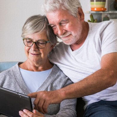 A man and woman in their sixties are sat together on a sofa. The woman, who is wearing a blue top and a grey cardigan, is looking at a tablet and the man, who is wearing a white t-shirt and blue jeans, has his arms around her pointing at the tablet. They are both smiling.