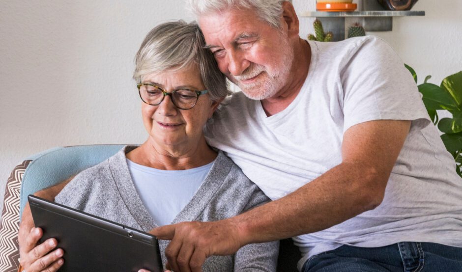 A man and woman in their sixties are sat together on a sofa. The woman, who is wearing a blue top and a grey cardigan, is looking at a tablet and the man, who is wearing a white t-shirt and blue jeans, has his arms around her pointing at the tablet. They are both smiling.