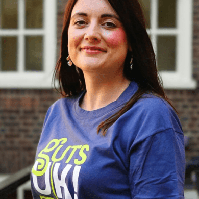 Amy has long dark hair. She is smiling at the camera and wearing a navy blue Guts UK t-shirt.