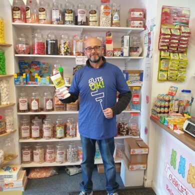 A person wearing a Guts UK Charity branded t-shirt is standing in a sweet shop, holding some sweets.