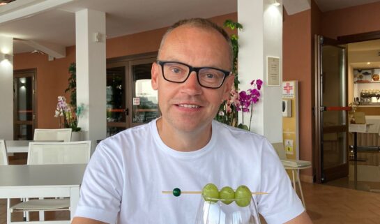 Jonald is sitting in a restaurant on holiday, smiling, with a glass of white wine in front of him