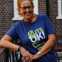 Suzanne is outside, she is standing up with her arm leaning on a railing. She is wearing a Guts UK branded t-shirt and glasses. Her hair is tied up. She is smiling at the camera.