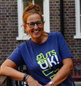 Suzanne is outside, she is standing up with her arm leaning on a railing. She is wearing a Guts UK branded t-shirt and glasses. Her hair is tied up. She is smiling at the camera.