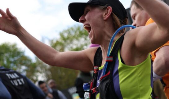 A person wearing a sports cap and a running vest has their hands up in the air, with a joyful expression on their face as they run amongst a crowd of runners.