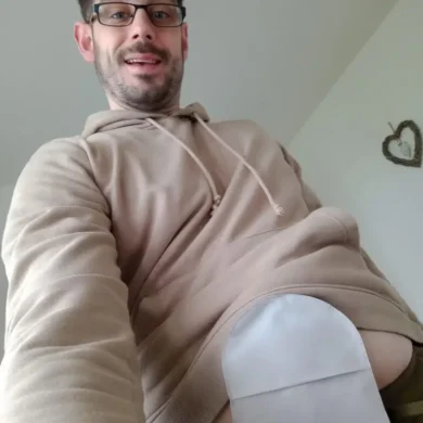 Ben is taking a selfie and is wearing a beige hoodie, black rimmed glasses and smiles at the camera. His ostomy bag is on show.