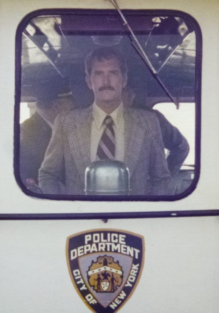 Tony is on a police boat during his career in the police force in Bermuda. The photo is taken through the window and he is looking at the camera with a neutral expression.