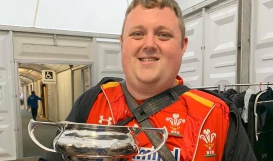 Gareth is wearing a red football shirt and is smiling at the camera, holding a trophy.
