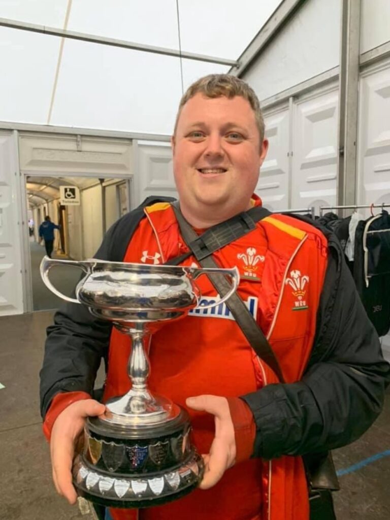Gareth is wearing a red football shirt and is smiling at the camera, holding a trophy.