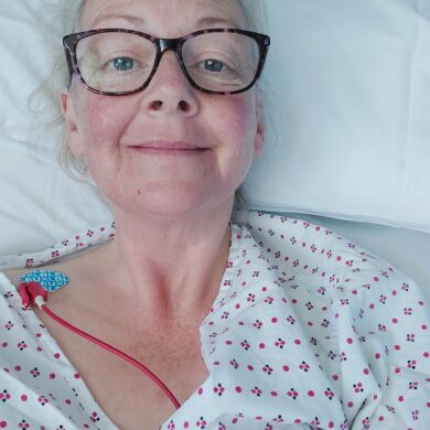 Karan is lying in a hospital bed. She is wearing a white patterned hospital gown and you can see a red tube running into her shoulder, which is covered by a plaster. She has short, light coloured hair and is wearing glasses with a dark frame. She is smiling at the camera.