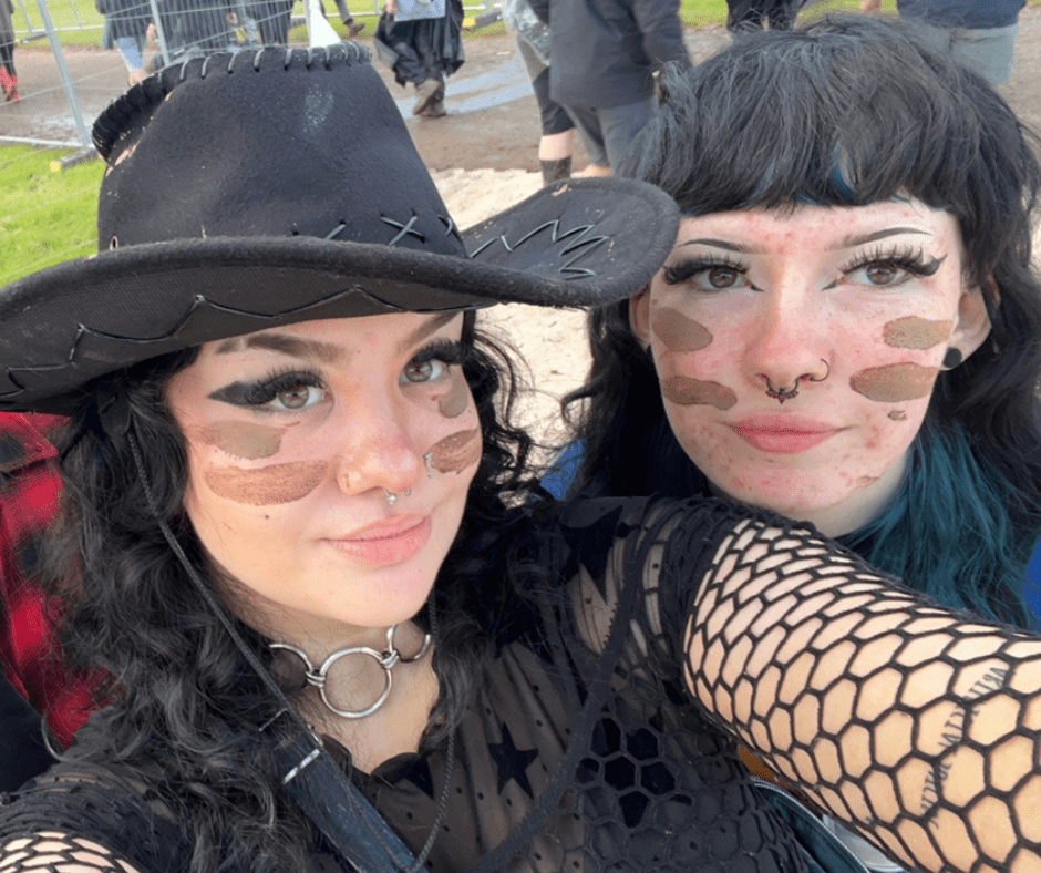 Rachel (left) wears a black cowboy hat, with fishnet style sleeves and a top and has black, long, curly hair. She is smiling at the camera and has two stripes of brown face paint on each of her cheeks. She is with her friend at a music festival.