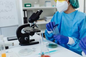 A scientist wearing gloves, scrubs, a mask and a scrub cap holding test tubes in a laboratory. There is a microscope and petri dish in front of him.