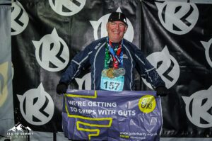 Cornelius is smiling with several medals on his neck. He is holding a Guts UK banner.