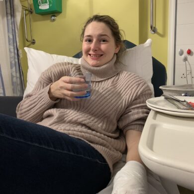 Sian is wearing a hazelnut jumper with black leggings and smiling at the camera. She is sat on a hospital bed with her left arm bandaged up and holding a glass of water on her right hand.