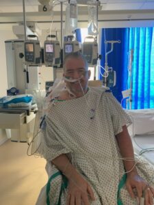 Cornelius is wearing a hospital gown in the hospital and sits in front of four machines that have tubes connected to his body. He is smiling at the camera.